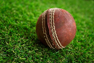 A worn and battered red leather cricket balls sits on the green turf