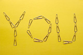 The word “YOU” formed with paper clips on a blank bright yellow paper