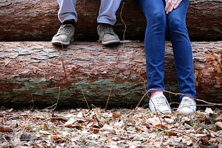 Two people sit on large, fallen trees, only their sneakers visible against the wood with little space between them.