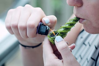 Person holding a green weed pipe in the mouth while lighting cannabis leaves in it.