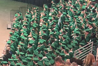 A group of graduates sitting in during commencement — all in green cap and gown.