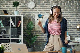 a woman wearing headphones, is in an office, standing up from the table where her computer is sitting, she is dancing to the music playing through her headphones
