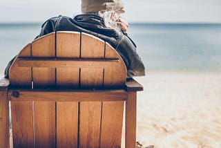 Sitting on a wooden deck chair looking out to sea with chin in hand, waiting.