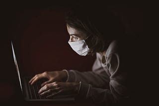 A woman wearing a mask typing on a laptop in the dark