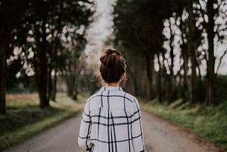 A woman in a chequered shirt and her tied in a bun walking away into a road that is lined both sides with trees.