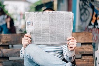 A person sites on a park bench reading a newspaper, holding the paper so that it blocks their entire face.