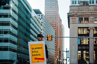 Photo of a man stood on a street in a city, holding a yellow sign which reads “slavery still exists”.