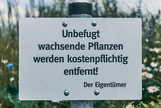 White sign on metal pole in flowery summer garden, in German, reading “Unbefugt wachsende Pflanzen werden kostenpflichtig entfernt! Der Eigentümer”