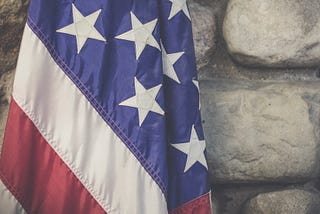 A close up of a partially-folded American flag, with the creases in its canvas visible, in front of blurred gray stone wall.