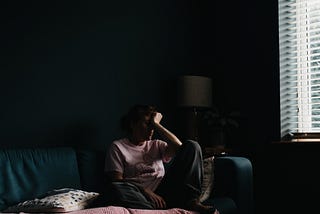 A woman sits in the dark alone on her couch, looking depressed and burned out. Many of us who feel like this struggle with self-care, but KC Davis says that’s okay, care tasks are morally neutral.