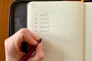 A journal in a gray case is open to a fresh page. A left hand holding a maroon pen has written “1. Why? 2. Why? 3. Why? 4. Why? 5. Why?”