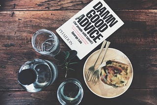 A table with a book Damn Good Advice, a wine glass, a coffee cup and a plate with some food and cutlery on it