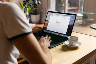 A man sits facing his laptop and typing. The laptop shows a browser with Google’s search bar.