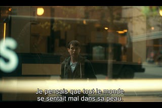 Capture d’écran de la série Umbrella Academy. On y voit Viktor, qui vient tout juste d’entamer sa transition et de faire son coming out à ses adelphes. Il observe son reflet dans une vitrine en expliquant des choses à sa soeur. “Je pensais que tout le monde se sentait mal dans sa peau”.