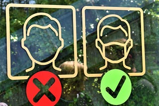 Closeup of decals on what appears to be a window of a business. There are some blurry reflections in the glass of people talking. The decals show two square yellow icons side by side. On the left, a big “X” is beneath the icon of a simple stick-figure-esque outline of a maskless head. On the right, a big checkmark is beneath the same stick figure outline of a head, but this one is wearing a face mask.