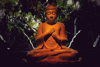 Image shows a clay statue of theBuddha, hands across his heart against a background of greenery.