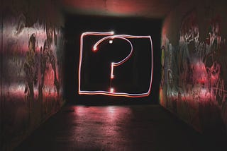 A red big neon-like question mark in the middle of a dark corridor. The sign casts some white, reddish, and greenish reflection on the corridor’s floor, ceiling, and the graffitied walls.