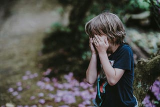 Young boy in dark blue t-shirt is covering his face as if crying. There are blurred pink/purple flowers in the background.