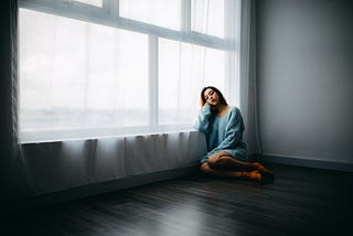 A woman is sitting on the floor near the window of an empty apartment. Anthony Tran/Unsplash.