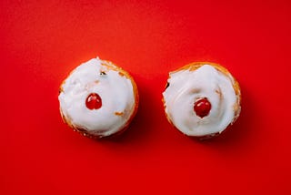 Two vanilla cupcakes with red cherries in the center, meant to look like breasts, set against a red backdrop.
