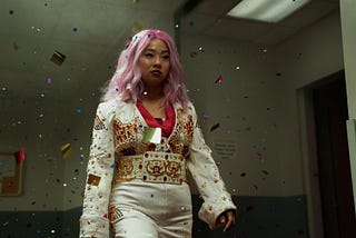 Jobu Tupaki, the villain of the movie Everything Everywhere All At Once, is walking in a hallway through a cloud of confetti. She has pink hair and is wearing a white jump suit that is bedazzled with ornate designs.