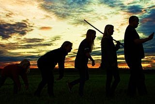 An image illustrating human evolution. A series of figures is seen in silhouette against a cloudy sunset. A crouched ape at the extreme left begins to rise, then is nearly upright and holding what seems to be a stone tool. The next figure appears to carry a spear or farm implement over its shoulder. In an ironic twist, the final figure on the right stares into a cell phone.