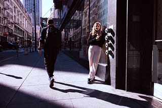 Blond lady standing in the sun, reading from her phone while a man dressed in black walks past her.