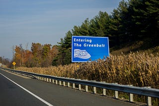 A road sign informs drivers they are entering the Greenbelt in Ontario. The highway is near a forest on a beautiful fall day.