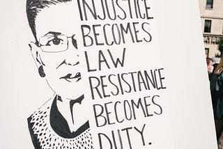 A line drawing of Supreme Court Justice Ruth Bader Ginsburg held by a demonstrator, and a quote”When Injustice becomes Law, Resistance becomes Duty.’’