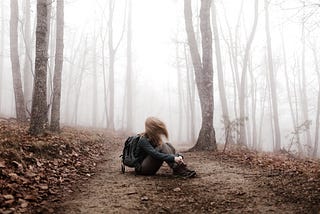 Winter forest, muted colors, girl with daypack sits on trail facing to the side; her hair blowing across covers her face