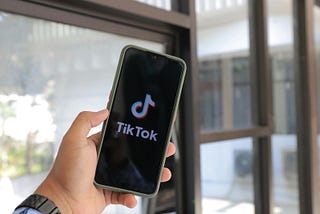 TikTok logo displayed on a smartphone screen held in hand, set against a bright background with window reflections.