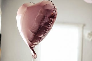 A pink, heart shaped balloon floating in a white walled room.