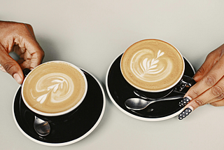 A close-up image of two hands holding black mugs filled with latte