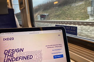 An open laptop in the foreground with the Interaction 23 website showing. The laptop is being used by someone on the train to Zürich, and you can see the countryside pass in the train window behind the computer.
