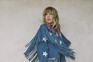 Taylor Swift looks coyly aside before a neutral background, wearing a fringey denim top with star patches