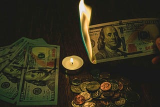 A hand holding a burning $100 bill above a pile of coins and other bills next to a lit candle.
