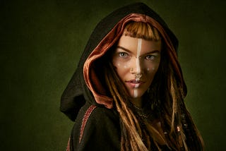 A young woman with fair skin, piercing amber eyes, long brown hair, a dark hooded cloak, and a white line down the middle of her face