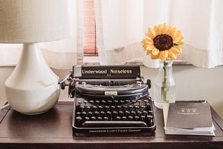 A typewriter, a white lamp, a sunflower in a vase, and a notebook