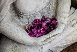 Purple flower petals are found gently cradled in the arms of a statue.