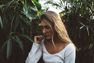 A woman in a white blouse lookng down. Backdrop is foliage.