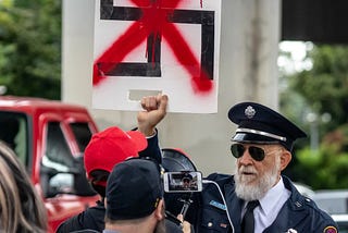 A man in a uniform holds up a sign with a swastika, crossed out in red paint, as a crew films him