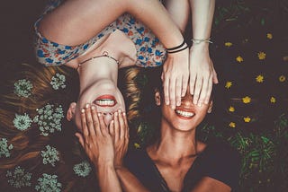 Two girls are lying next to each other. Their heads are next to each other but their bodies are in opposite directions. They are covering each other’s eyes with their hands and smiling.