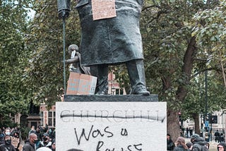 An image of a group of people in protest against a Winston Churchill statue that has been spray painted with words to read “Churchill was a racist”. The statue holds up a sign that reads “Black Lives Matter”