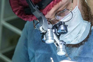 A close-up of a surgeon wearing magnifying loupes, a headlamp, surgical mask, and red cap while performing a procedure. The focused lighting highlights the precision tools and the surgeon’s intense concentration.
