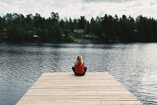 A blonde woman in a red sweater sitting cross-legged on a pier, overlooking a lake