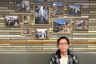 A young woman wearing glasses and a black and white plaid shirt sits at a wooden table in a restaurant.