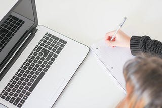 An individual with brown hair is writing her ideas on a notepad while looking at her laptop.