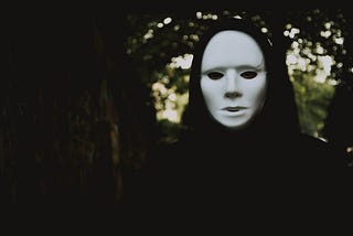 A cloaked figure in a plain white mask covering the face, intended to be symbolic of an unknown threat