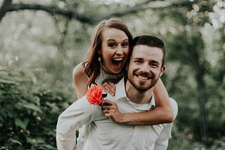 A picture of a happy couple, a man and a woman. The woman has jumped up onto her husband’s back and is holding a rose.