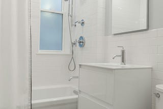 An image of a bathroom showing a shower unit, a sink and a toilet, all in white with white tiles on the wall.